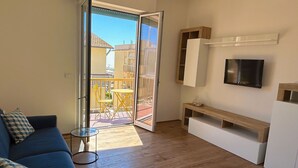 Una sala de estar moderna con un sofá, un televisor y un ventanal corredero que da a un patio exterior con sillas amarillas.