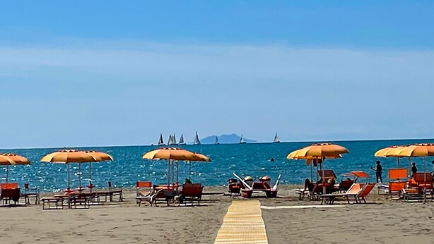 Una playa con hileras de sombrillas naranjas y tumbonas, un paseo marítimo de madera, y veleros a la distancia.