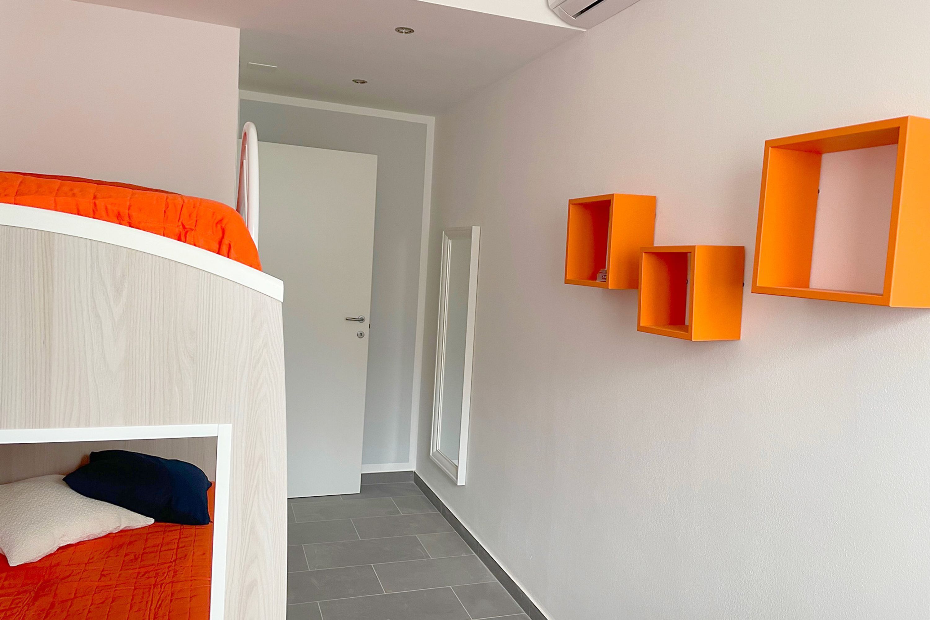 Et moderne værelse med køjeseng, en dør der fører til et andet værelse, og orange væghængte hylder.