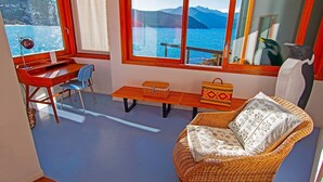 Une pièce comprenant un bureau, une chaise et un fauteuil en osier avec un coussin. Il y a une fenêtre donnant sur la mer et les montagnes.