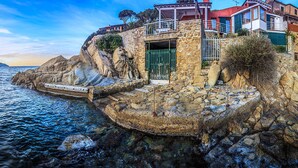 Une maison en pierre située sur la côte, dotée d’un balcon donnant sur la mer, d’un escalier en pierre menant à l’entrée et offrant une vue dégagée sur l’océan.