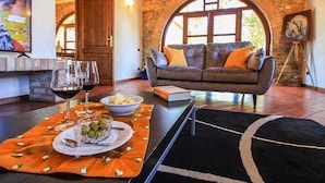 A cozy living room with a stone wall, a wooden door, a grey sofa with orange cushions, a black rug, and a table with a glass bowl of olives and wine glasses. - Villa Campo Valerio con Grande Piscina (Scarlino)