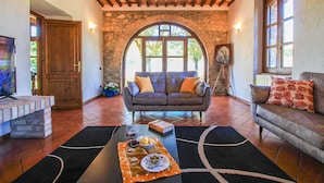 A cozy living room with a stone archway, a grey sofa with cushions, a wooden coffee table with a placemat, and a television on a stand. - Villa Campo Valerio con Grande Piscina (Scarlino)