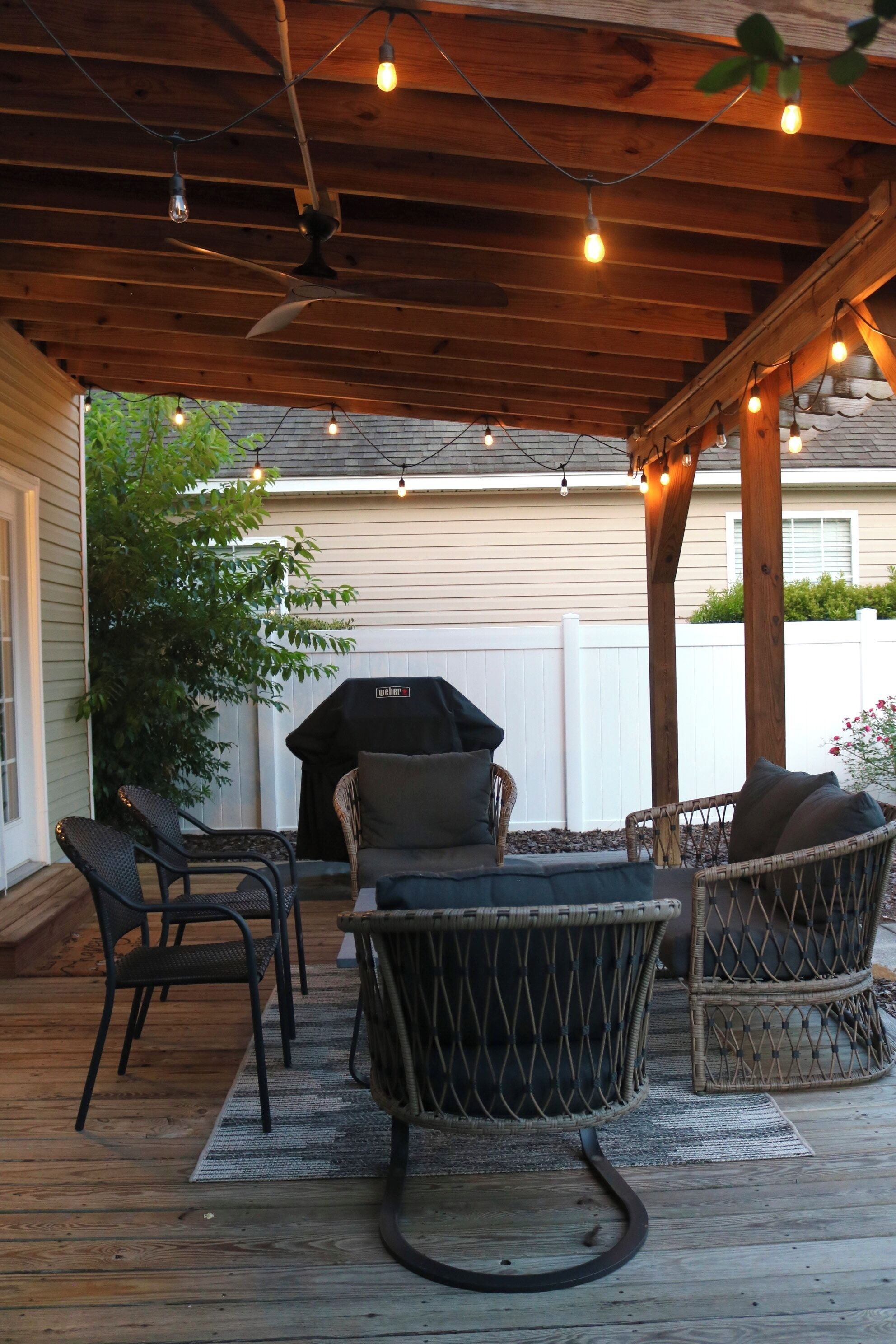 Un patio techado con mobiliario de mimbre, ventilador de techo, luces colgantes y una parrilla.