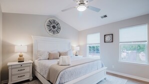 A bedroom with a large bed, a ceiling fan, a nightstand with a lamp, and a window with blinds.
