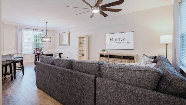 A modern living room with a grey sectional sofa, a ceiling fan, a dining area with a table and chairs, and a kitchen area with a wooden cabinet. - Welcome to the Lowcountry Lodge - cornhole, beach chairs, walk to town (Bluffton)