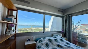 A bedroom with a large window, a bed with a peacock feather pattern, a wooden shelf, and a view of the sea.