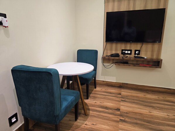 A room with a round white table, two blue chairs, a flat-screen TV mounted on a wooden shelf, and a power outlet with a phone and remote control.