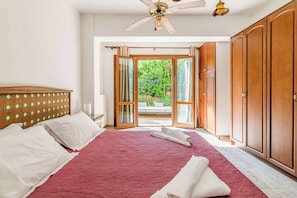 Un dormitorio con una cama grande, un ventilador de techo y vistas a un jardín a través de puertas abiertas.