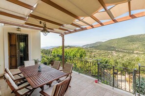 A balcony with a wooden table and chairs, a potted plant, and a view of a green landscape with hills. - Odyssey's Palace - Escape in Mythical Ithaca (Platrithias)