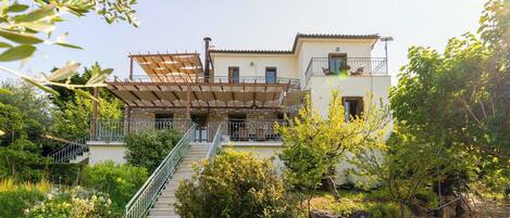 A two-story house with a staircase leading to a balcony, surrounded by greenery and a stone wall.