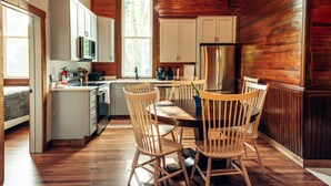 Een kleine keuken met houten vloer, witte kastjes, een roestvrijstalen koelkast, en een eethoek met houten stoelen.