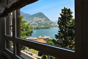 A view from a window overlooking a lake with buildings on a hillside and a tree in the foreground. - Luxury Lake View Motta Apartment in Lugano (Lugano)