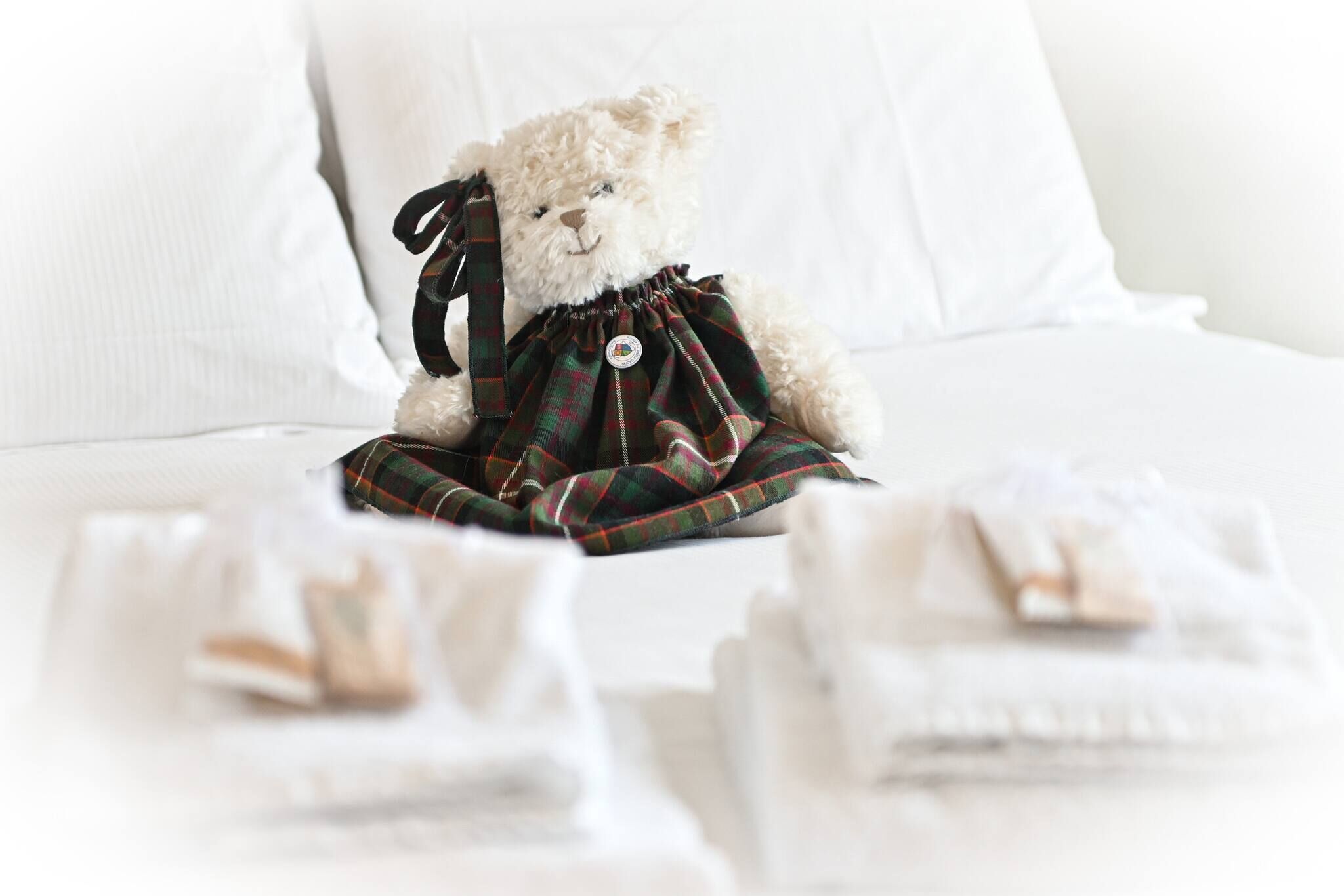 A plush toy in a plaid dress sitting on a bed with neatly folded white linens.