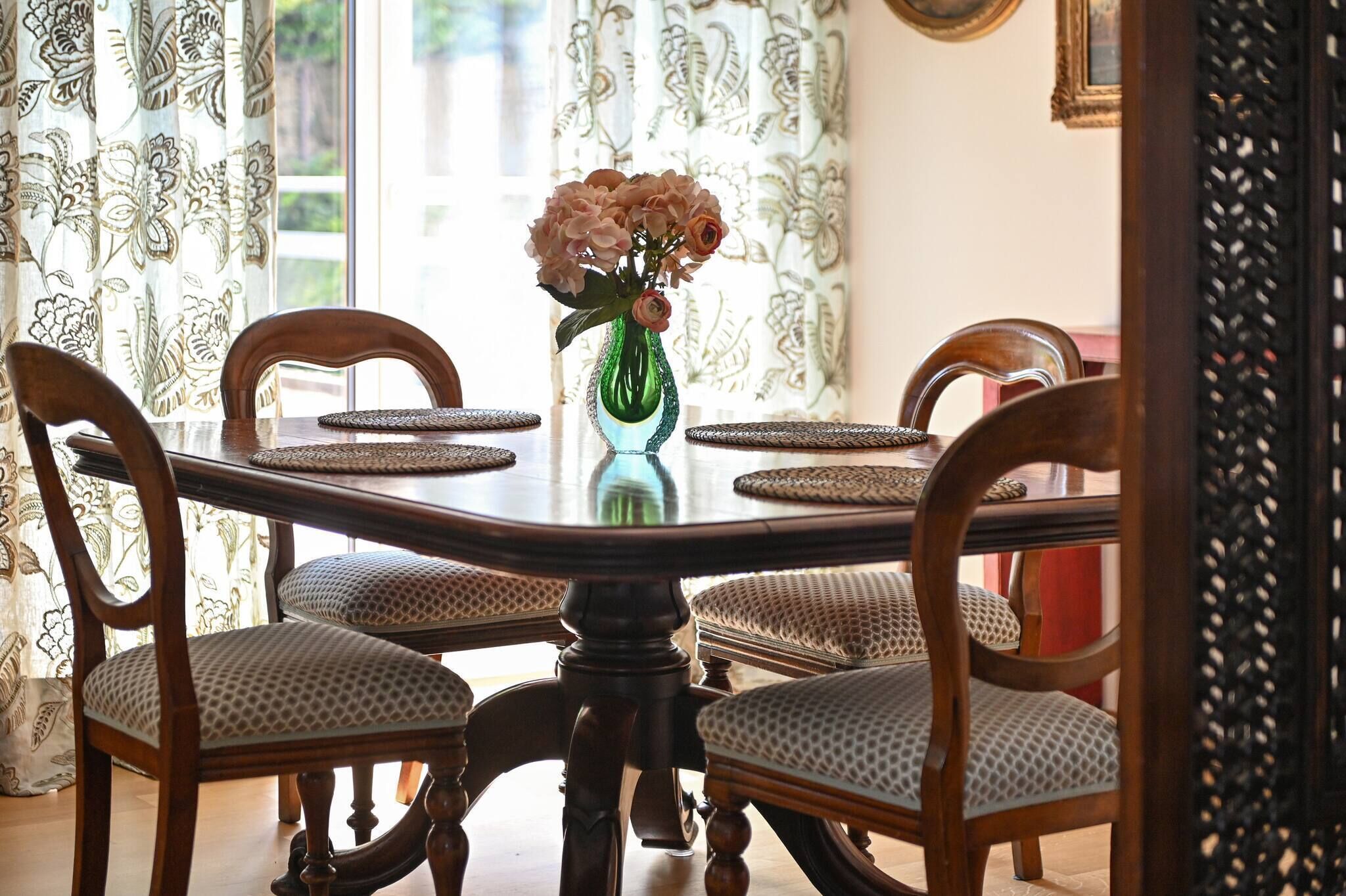 Una mesa de comedor con cuatro sillas, un jarrón verde con flores y cortinas con estampado.