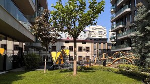 Une cour avec un arbre, des plantes à fleurs jaunes et du matériel de jeux pour enfants.