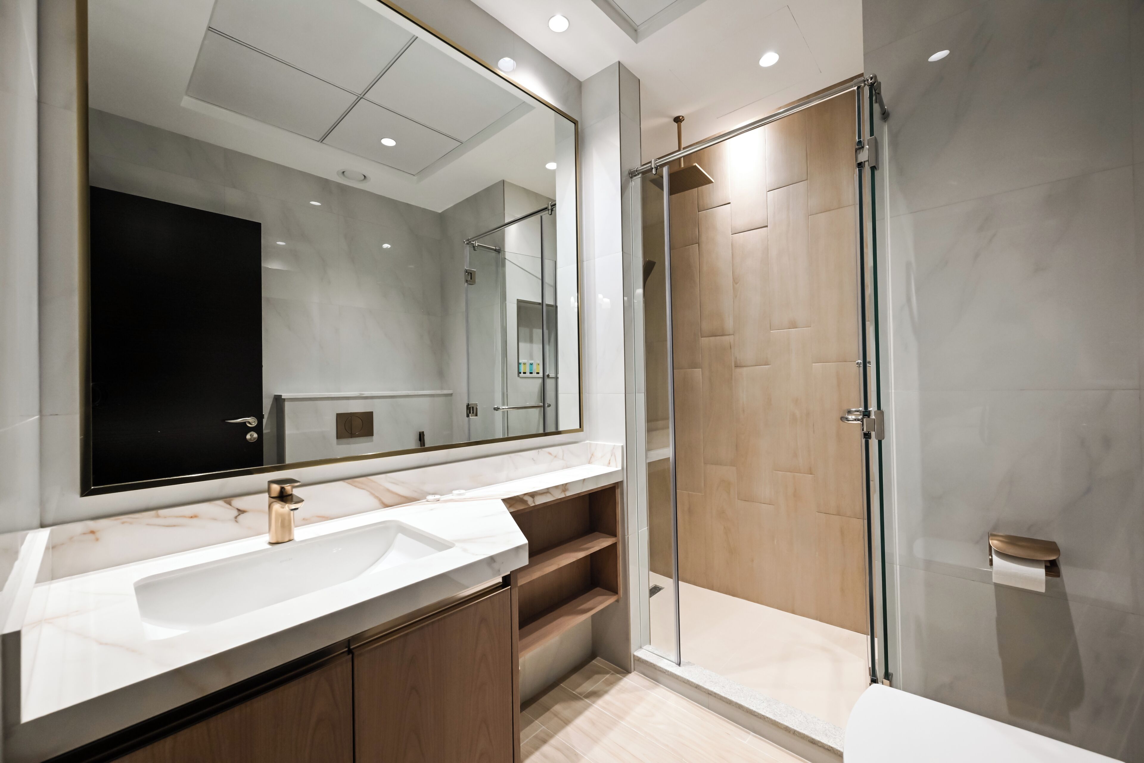 A modern bathroom with a large mirror, white marble countertop, wooden cabinets, a glass shower enclosure, and a toilet paper holder.