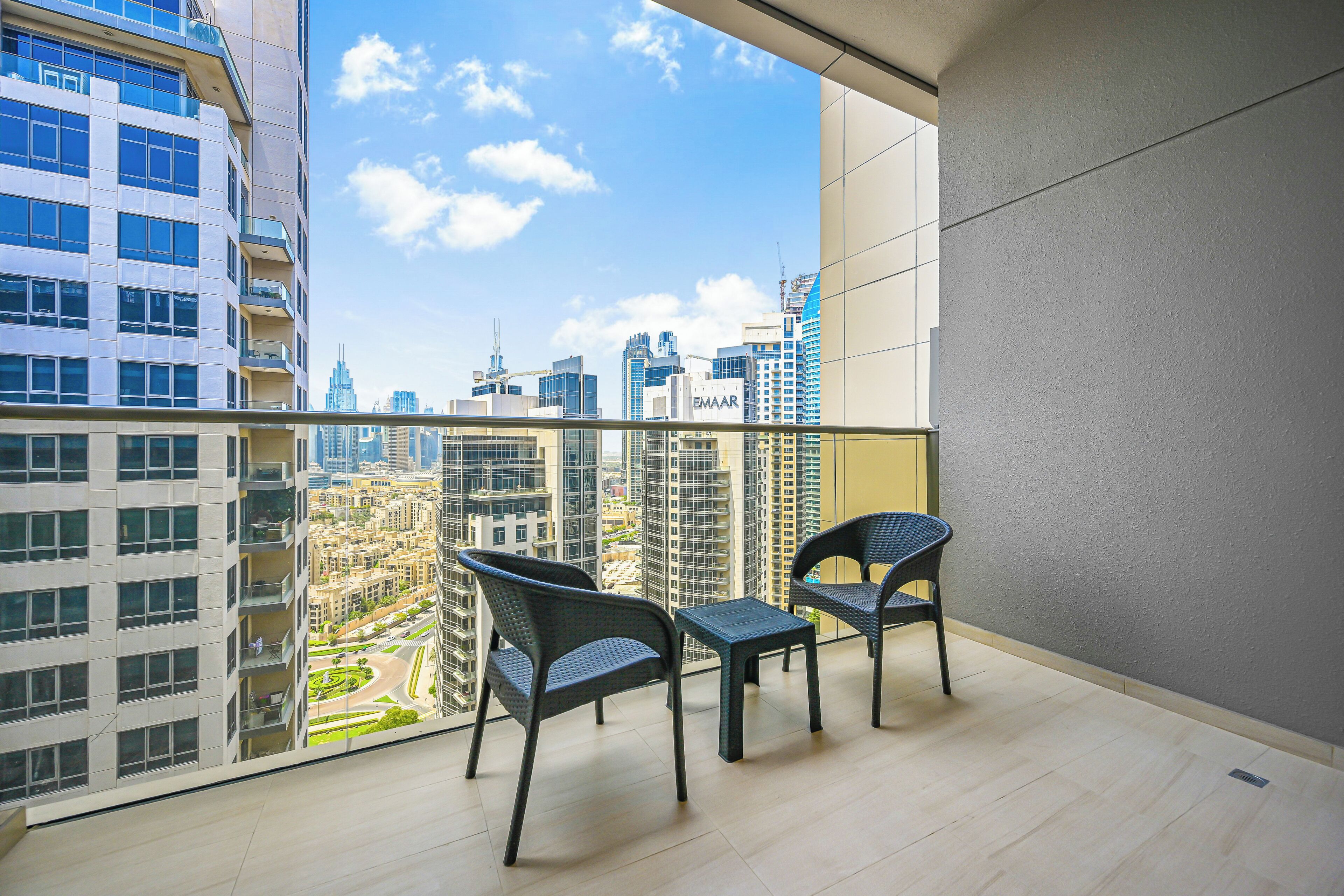 A balcony with two chairs and a small table, overlooking a cityscape with high-rise buildings and a clear sky.