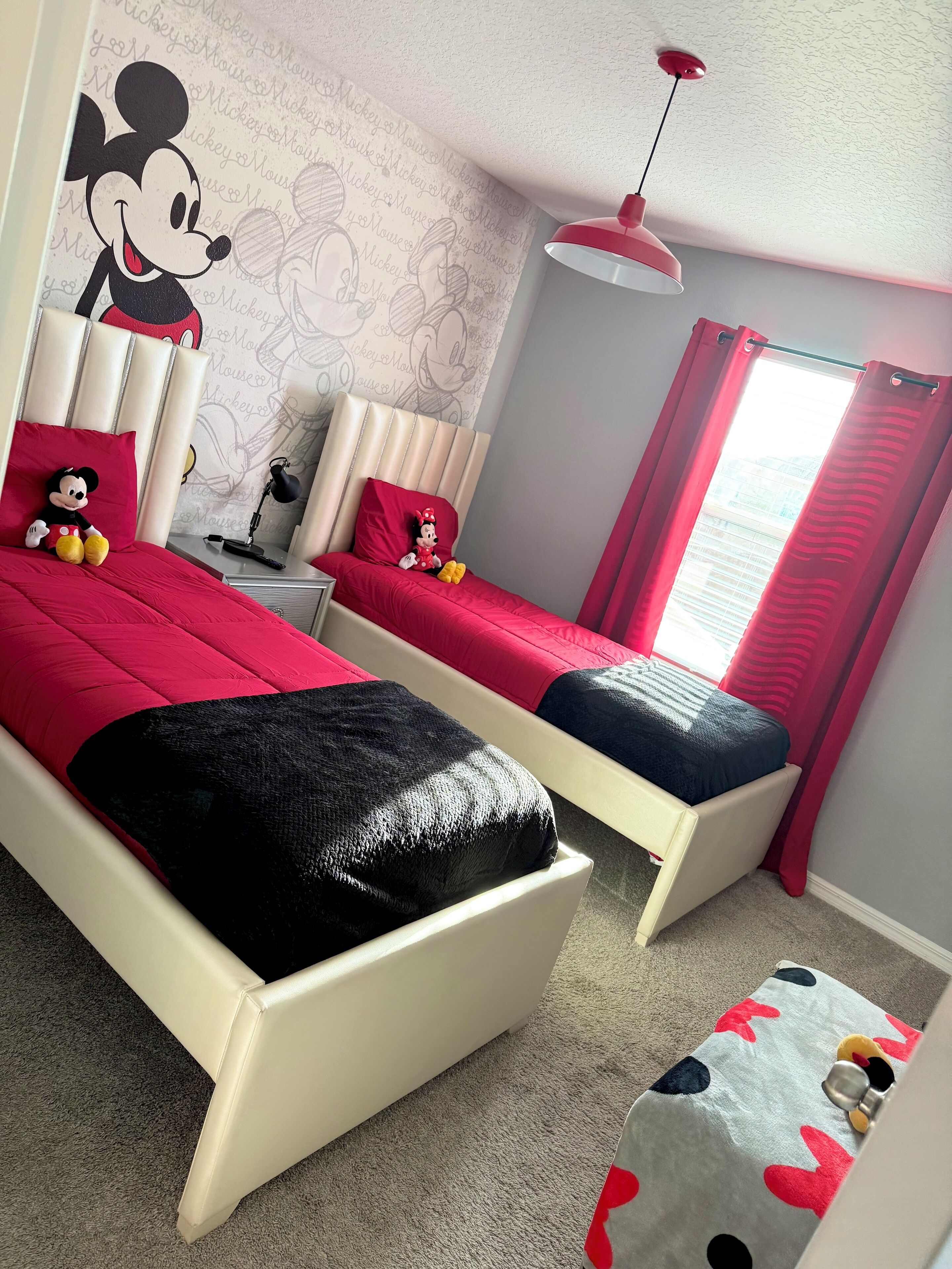 Una habitación con dos camas, cada una con cabecera temática de Mickey Mouse, un peluche de Mickey Mouse en una cama y una almohada de Mickey Mouse en la otra. Hay un escritorio con lámpara, una ventana con cortinas rojas y una lámpara de techo.