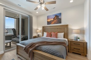 Ein Schlafzimmer mit einem Bett, einem Holzkopfteil, Nachttischen, einem Beistelltisch, einem Deckenventilator und einem großen Fenster.