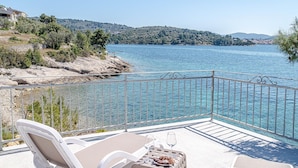 A balcony with a clear view of a lake, a chair, a table with a glass of wine, and a plate of pastries.