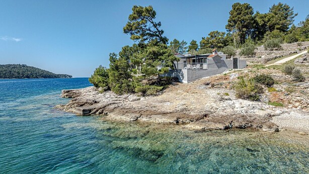 A coastal house with a rocky shoreline, clear blue sea, and a distant island.