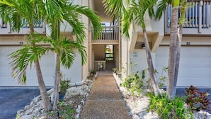 A pathway leading to a building with numbered garage doors, surrounded by palm trees and plants. - Siesta Beach Retreat 2 Bedroom Townhouse (Siesta Key)