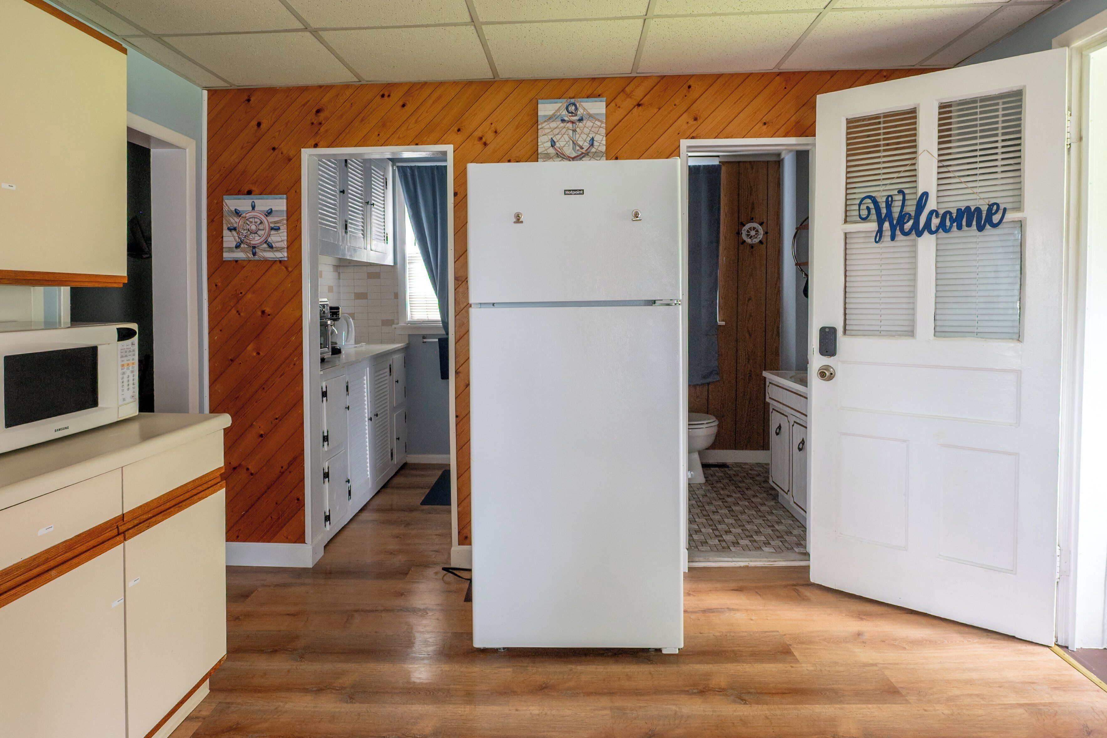 A compact kitchen with a white refrigerator, wooden panel walls, and a door with a 'Welcome' sign.