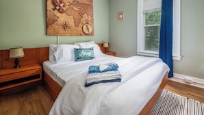 A neatly made bed with white linens, a world map artwork above it, a clock on the wall, and a window with blue curtains.