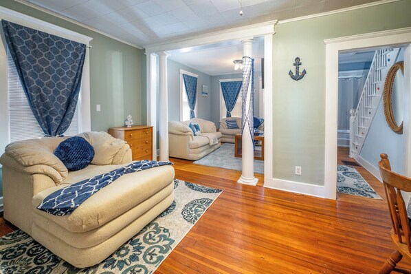Amplia sala de estar con un sofá color crema, almohadones estampados y piso de madera. Se ve una escalera que sube a un nivel superior, y un ventanal con cortinas azules.