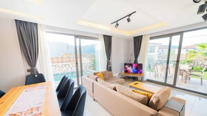 A modern living room with a sofa, a coffee table, and a TV. There is a dining table with chairs and a window with curtains. - Villa Meyra (Kas)