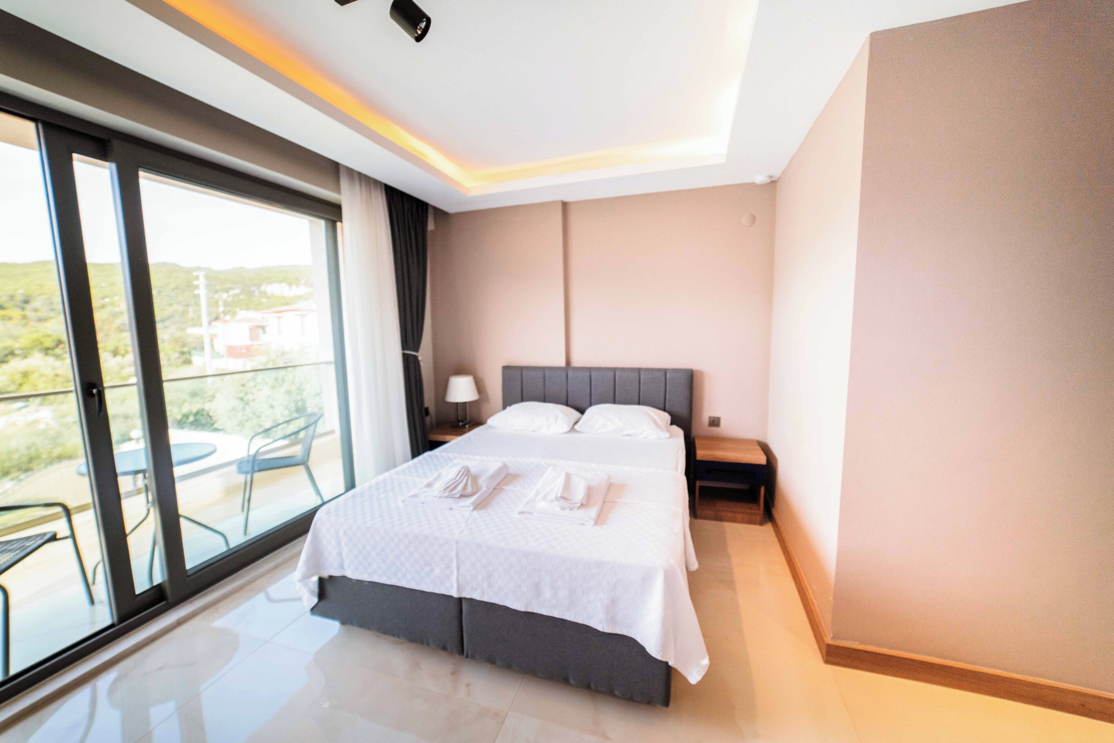 A modern hotel room with a large bed, a nightstand, a lamp, and a view of the outdoors through a sliding glass door.