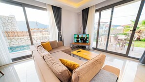 A modern living room with a beige sofa, a wooden coffee table, and a TV displaying a show. Large windows offer a view of a pool and outdoor furniture. - Villa Meyra (Kas)