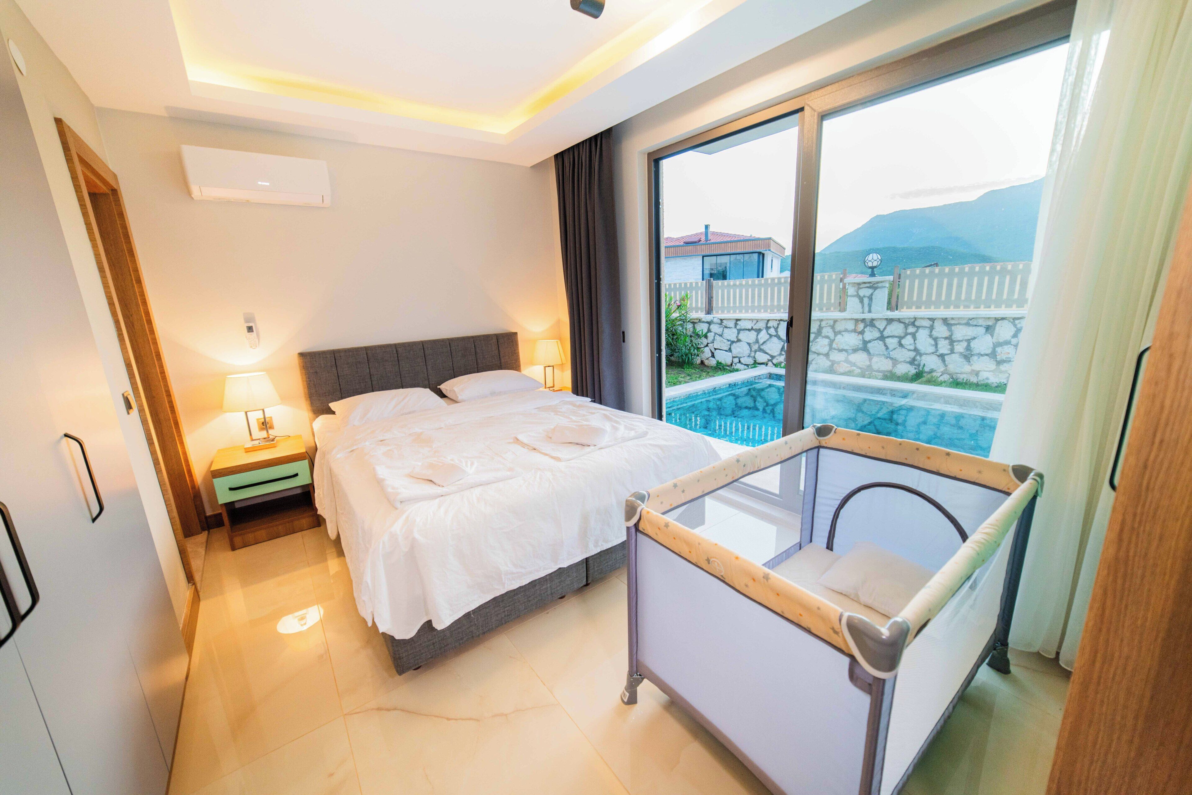 A hotel room with a bed, bedside table, lamp, and a view of a swimming pool and mountains.