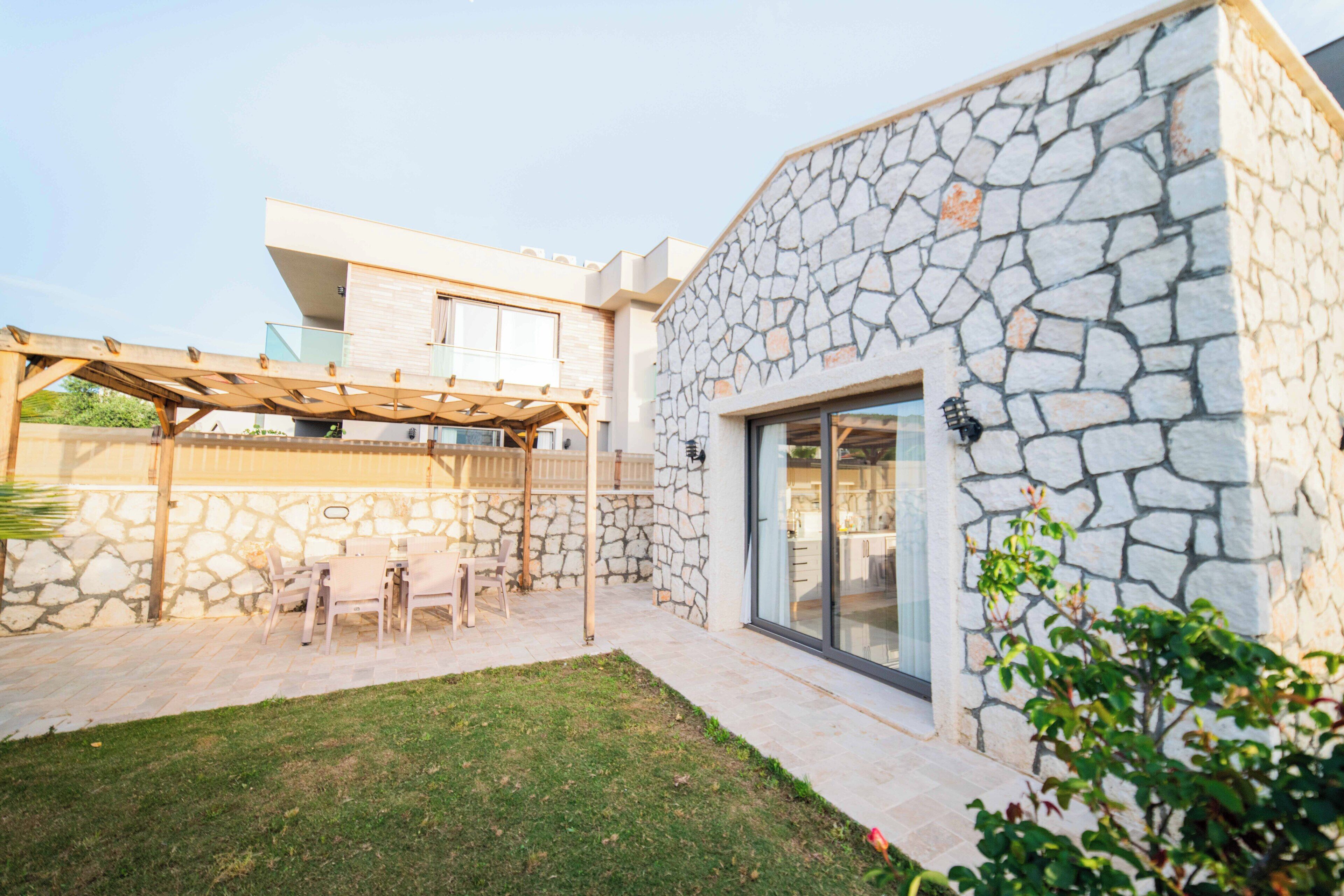 A modern house with a stone exterior, a covered patio with outdoor furniture, and a glass door leading to the interior.