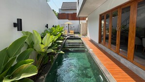 Un espace piscine extérieur moderne, doté d’une terrasse en bois, de grandes plantes vertes et d’un coin salon avec une table et des chaises.