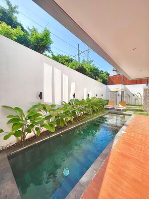 Une piscine dotée d’une terrasse en bois, entourée d’un mur blanc et d’une végétation luxuriante.