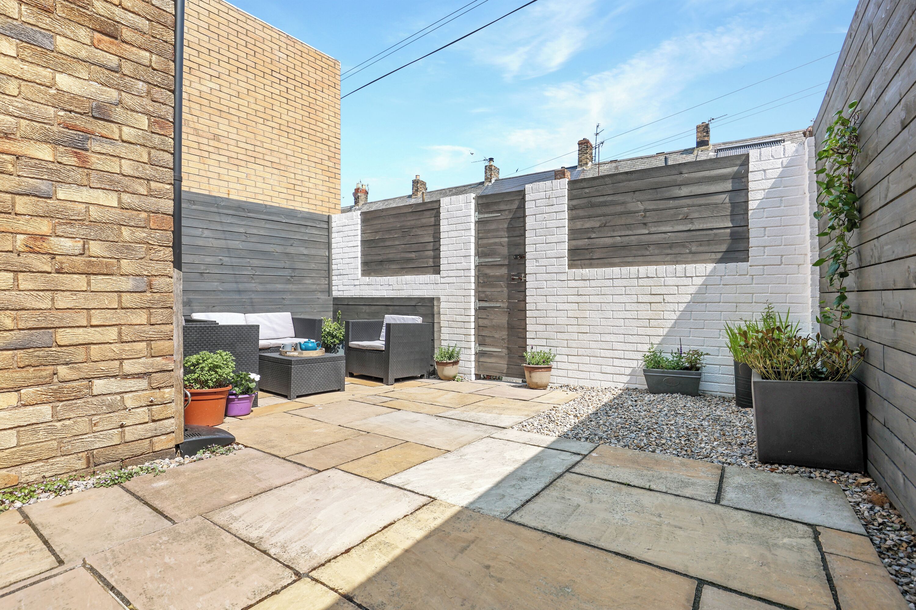 A courtyard with a brick wall, a white brick wall, and a grey wall. There are potted plants and a seating area with a table and chairs.