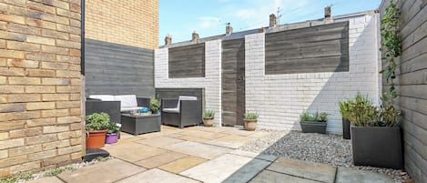 A courtyard with a brick wall, a white brick wall, and a grey wall. There are potted plants and a seating area with a table and chairs.