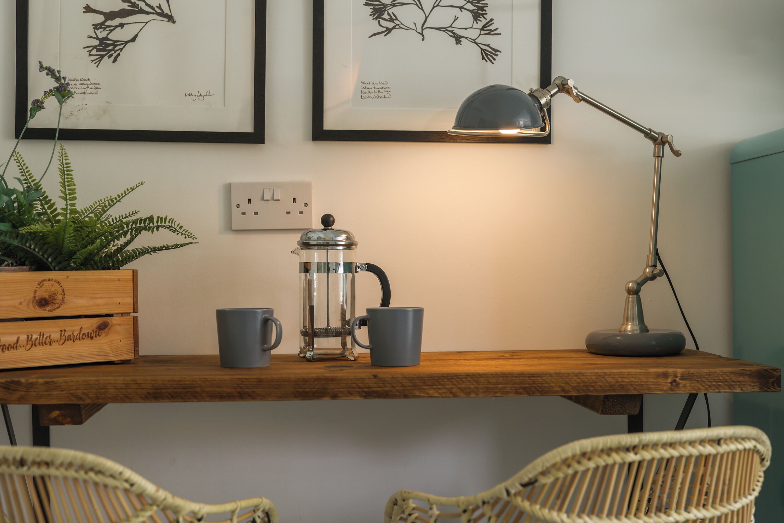 Una mesa de madera con una cafetera, dos tazas, una planta en maceta y una lámpara.