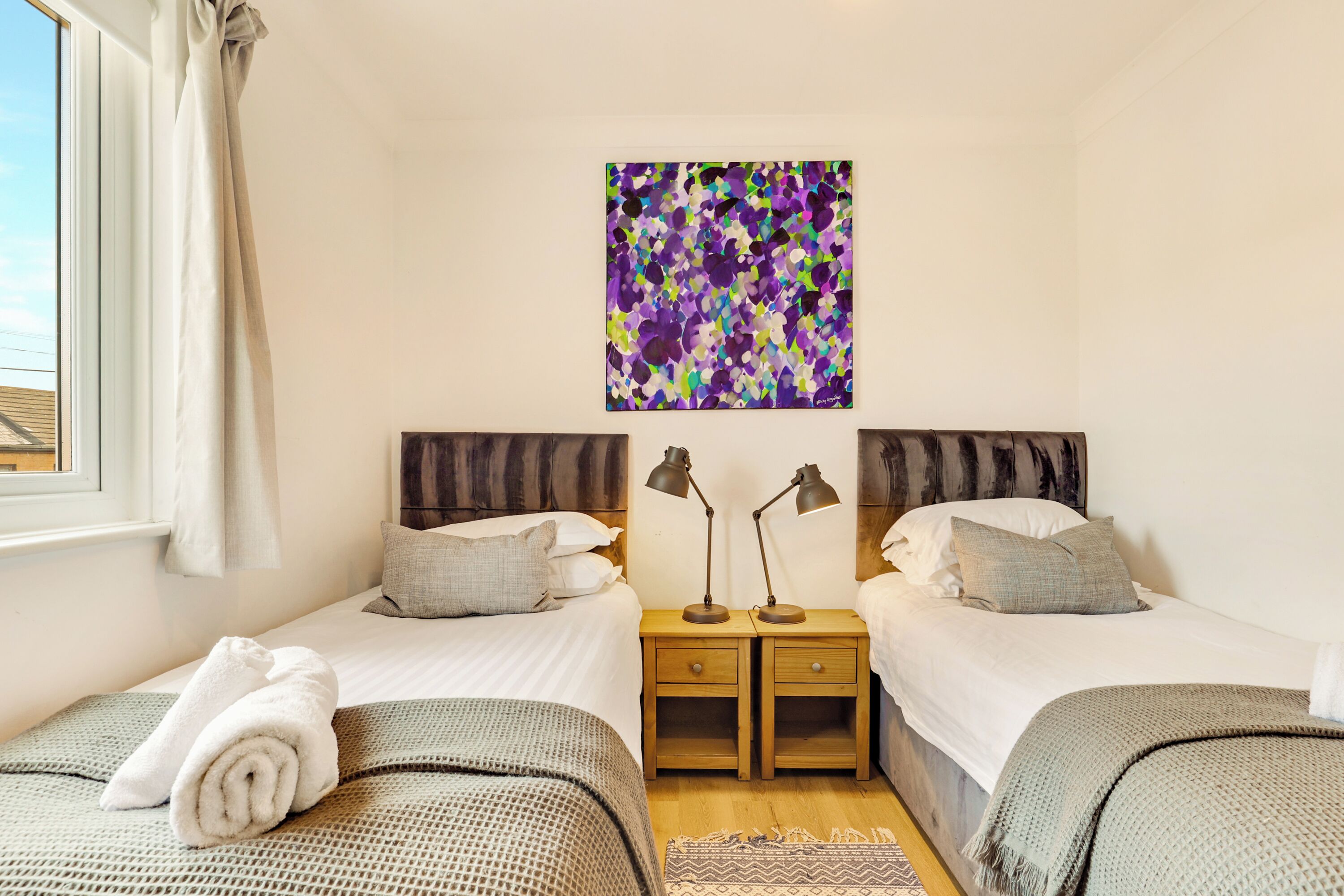 A twin room with two beds, a wooden nightstand with lamps, a colorful abstract painting, and a window with curtains.