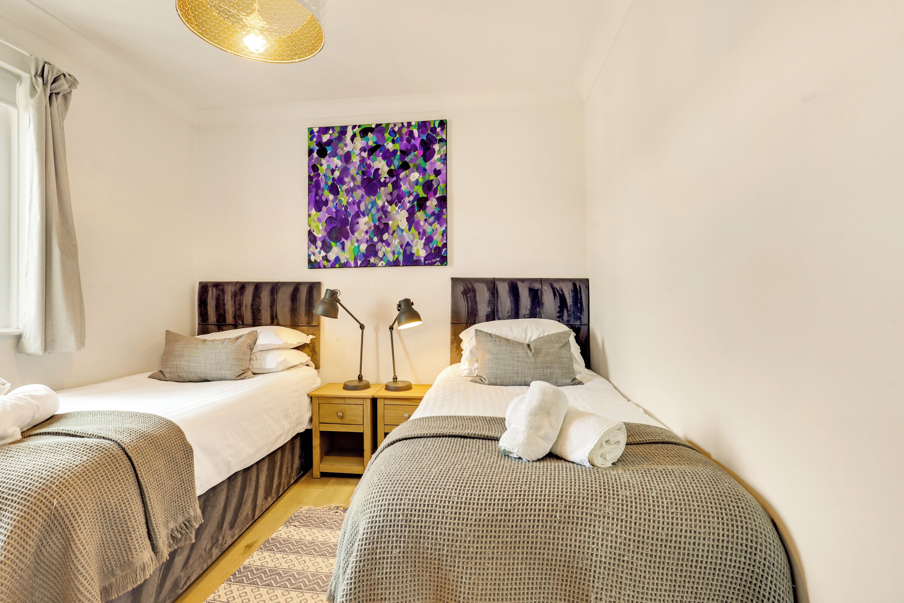 A bedroom with two single beds, a nightstand with lamps, a wall art piece, and a window with curtains.