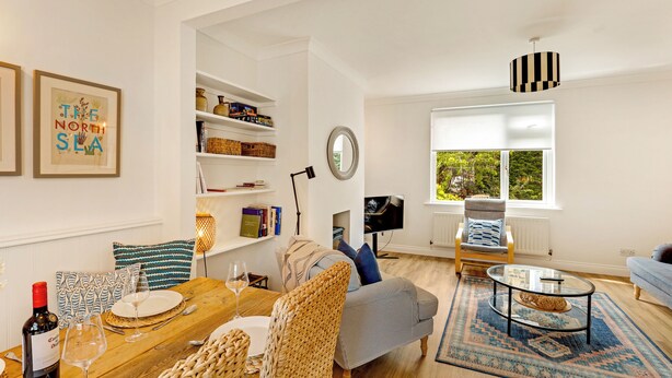A well-lit living room with a wooden dining table, a sofa, a TV, and a bookshelf. - Cherry Tree House - North East Escapes (Amble)