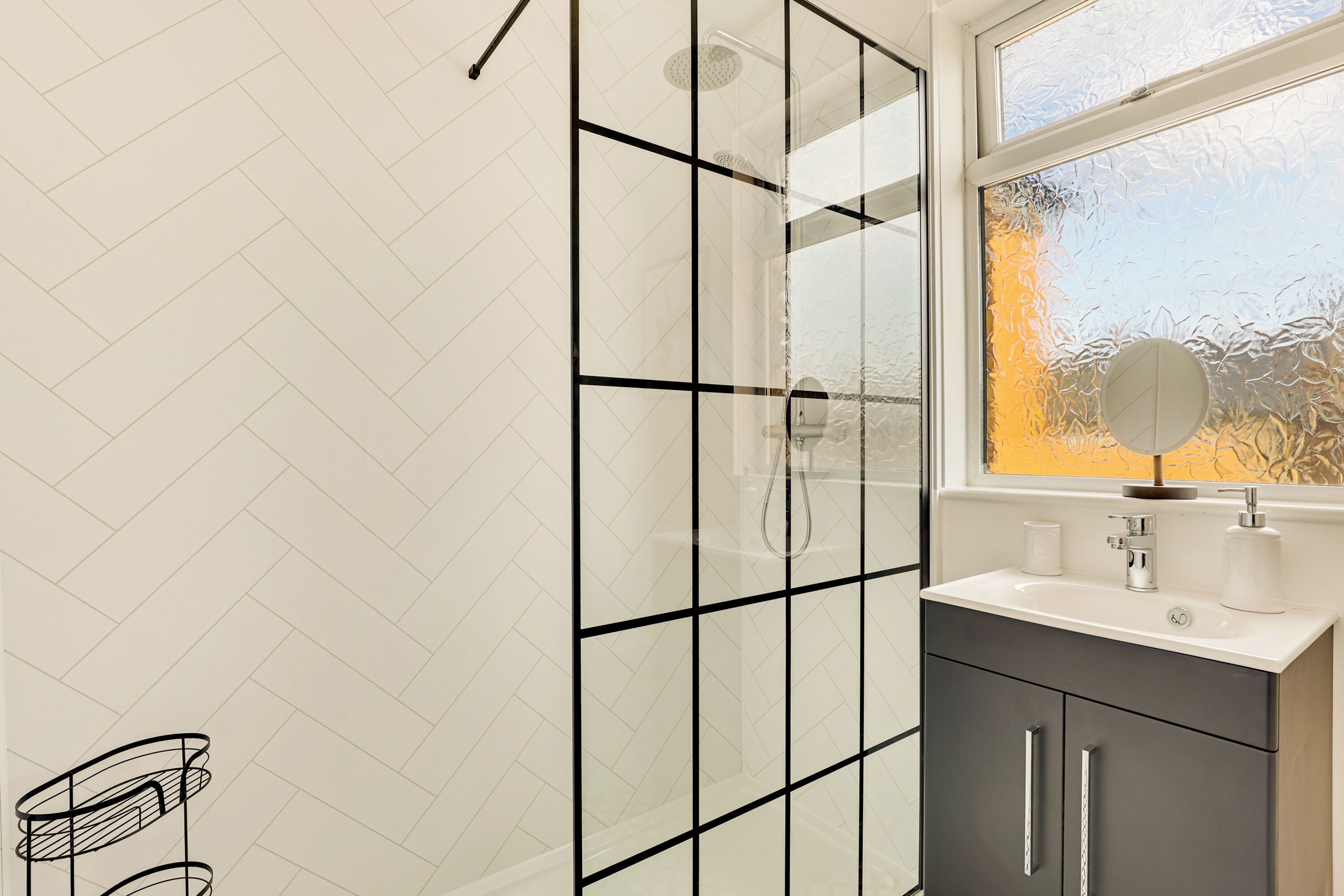 A modern bathroom with a glass shower enclosure, a white sink with a mirror, and a herringbone-patterned wall.