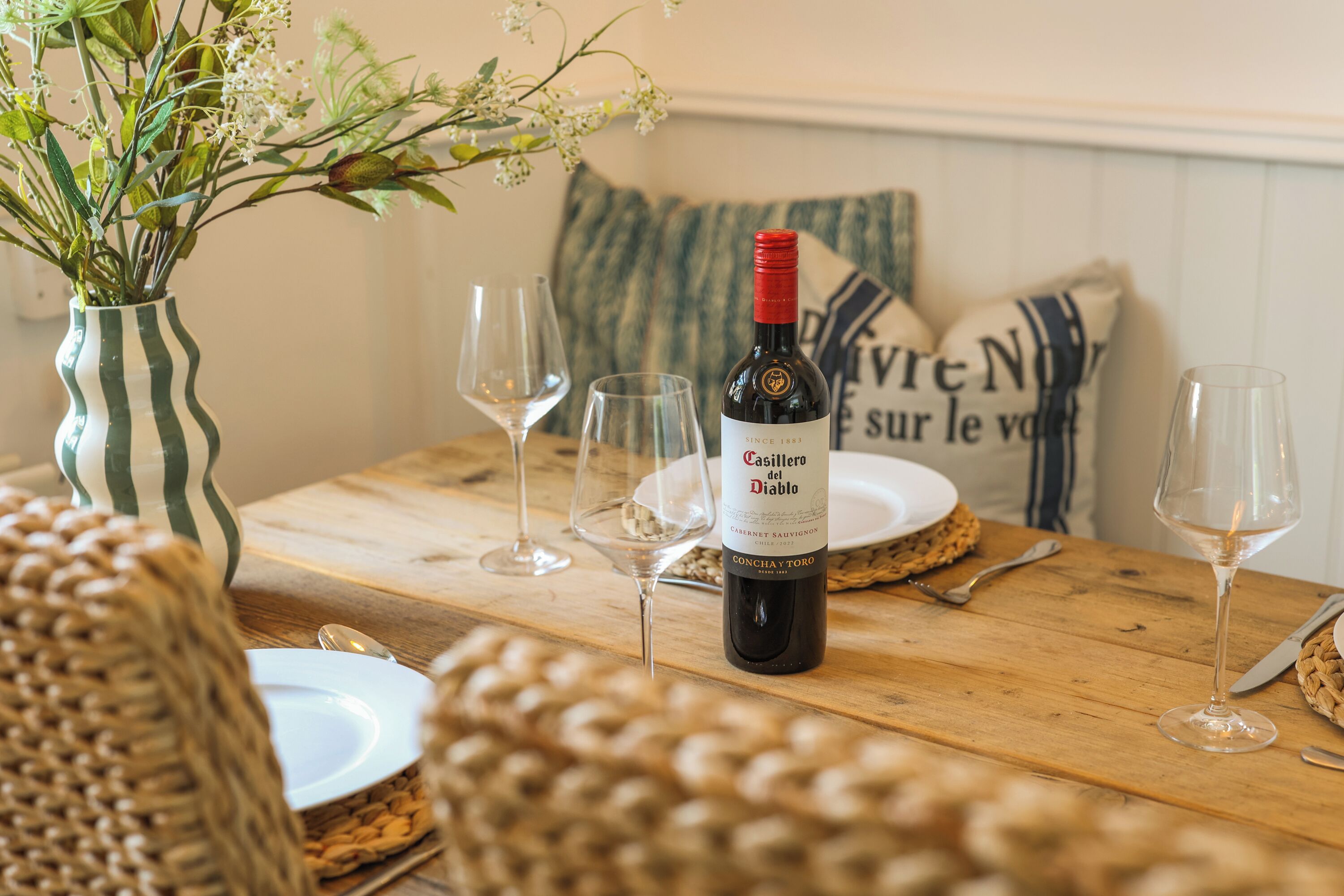 Een eettafel met een fles Casillero del Diablo wijn, twee lege wijnglazen en een vaas met bloemen.