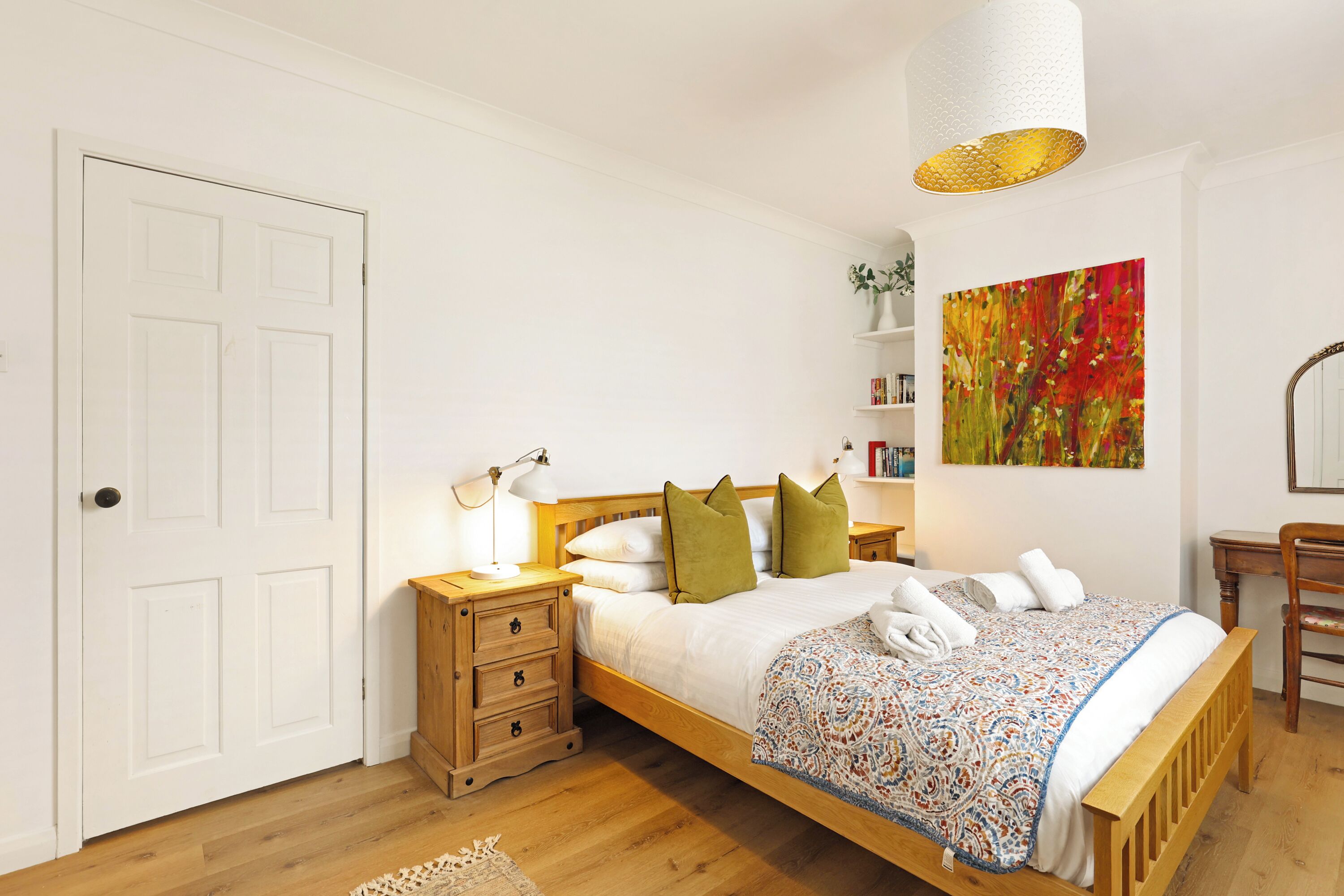 Un dormitorio con cama de madera, paredes blancas, una puerta blanca, una mesita de noche con lámpara y un cuadro abstracto de colores en la pared.