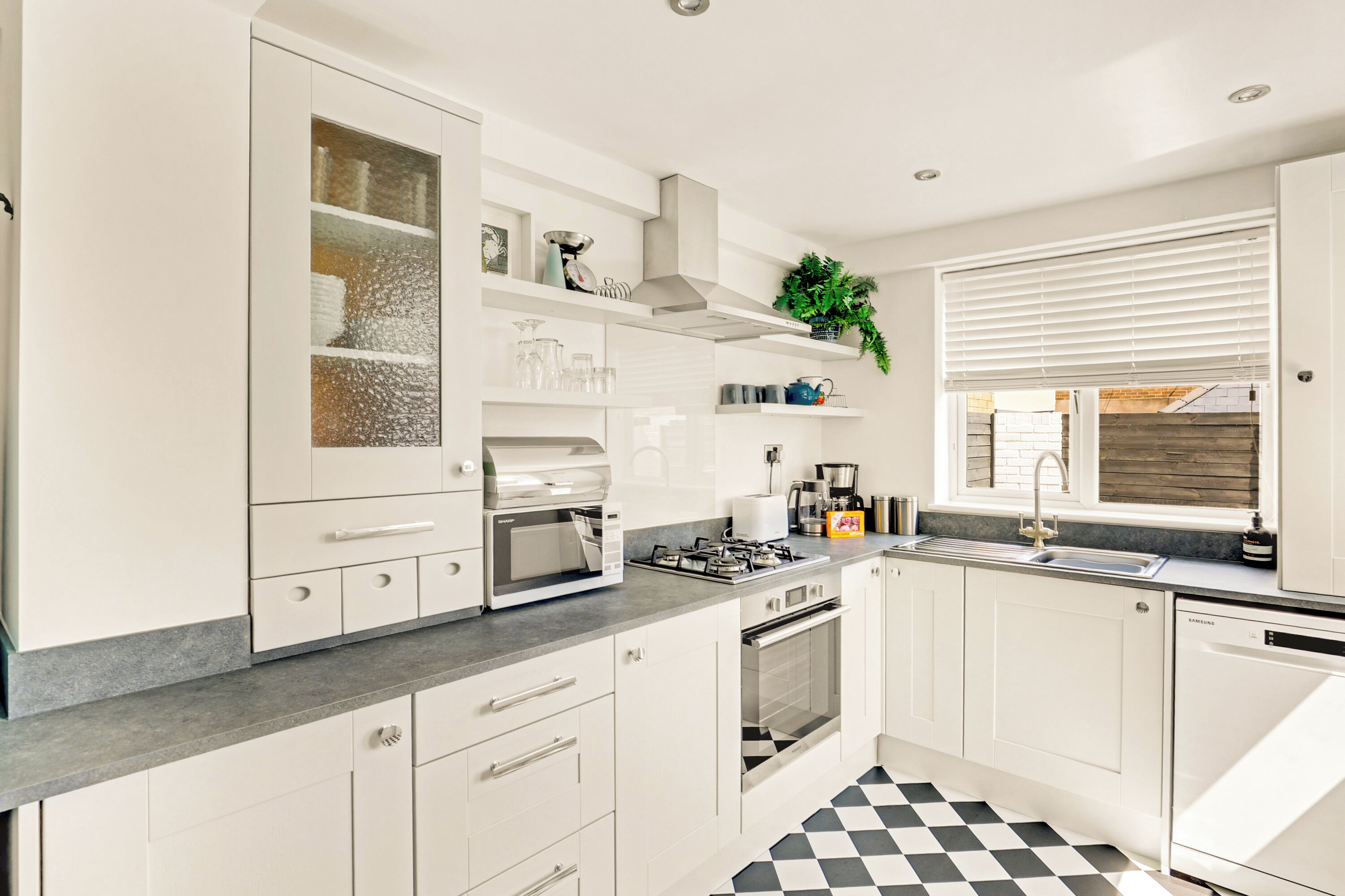 A modern kitchen with white cabinets, a black and white checkered floor, a built-in oven, and a sink with a window above it.