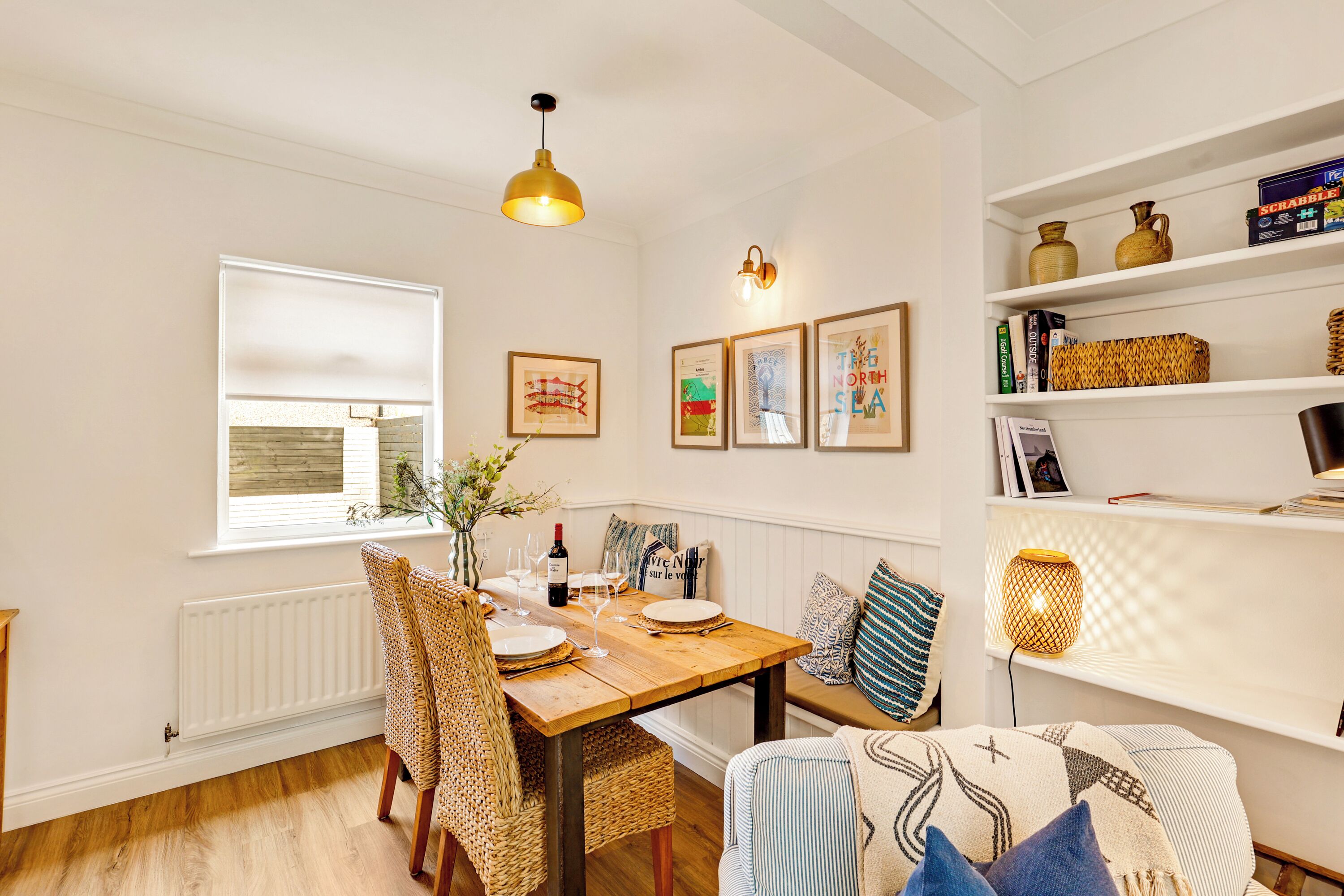 Un comedor con una mesa de madera dispuesta para cuatro personas, rodeada de sillas de mimbre. Hay obras de arte enmarcadas en la pared, una estantería con libros y objetos decorativos, y una lámpara colgante sobre la mesa.