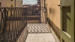 Un étroit couloir extérieur, délimité par une clôture métallique, doté d’un sol carrelé et bordé d’un bâtiment avec une porte verte sur le côté droit.