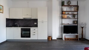 Una cocina moderna con armarios blancos, encimera negra y horno empotrado. Se aprecia un estante de madera con objetos decorativos y un televisor.